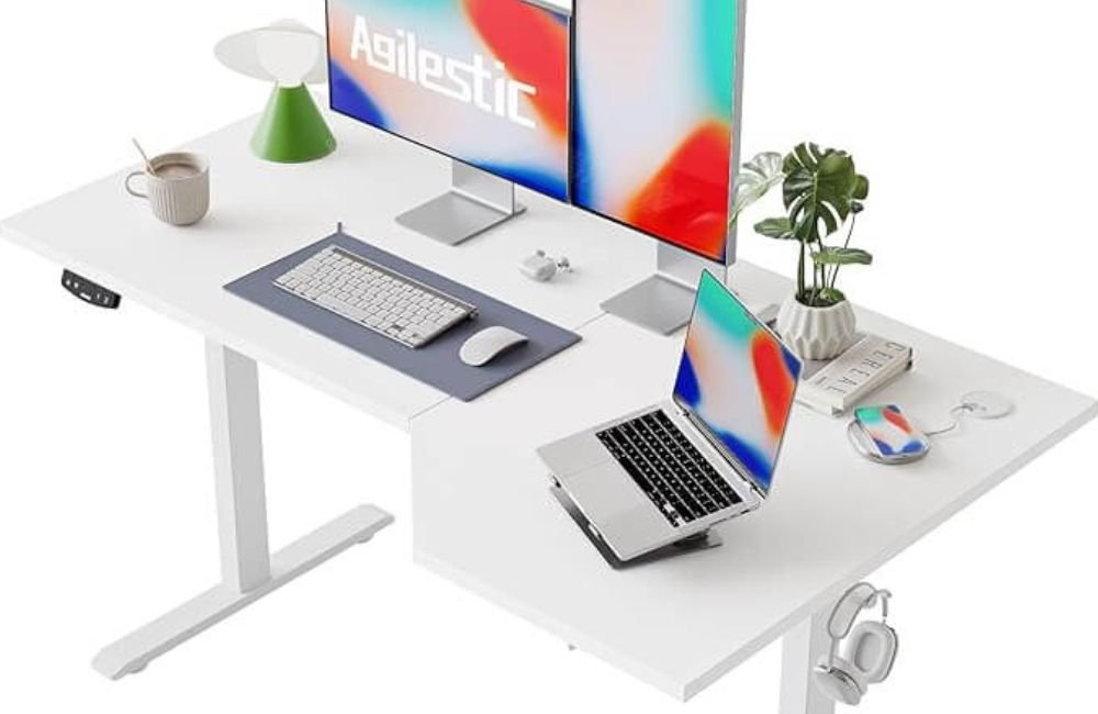 Agilestic electric standing desk in white finish with monitor displaying brand logo, laptop stand, and minimalist workspace accessories for productive remote work
